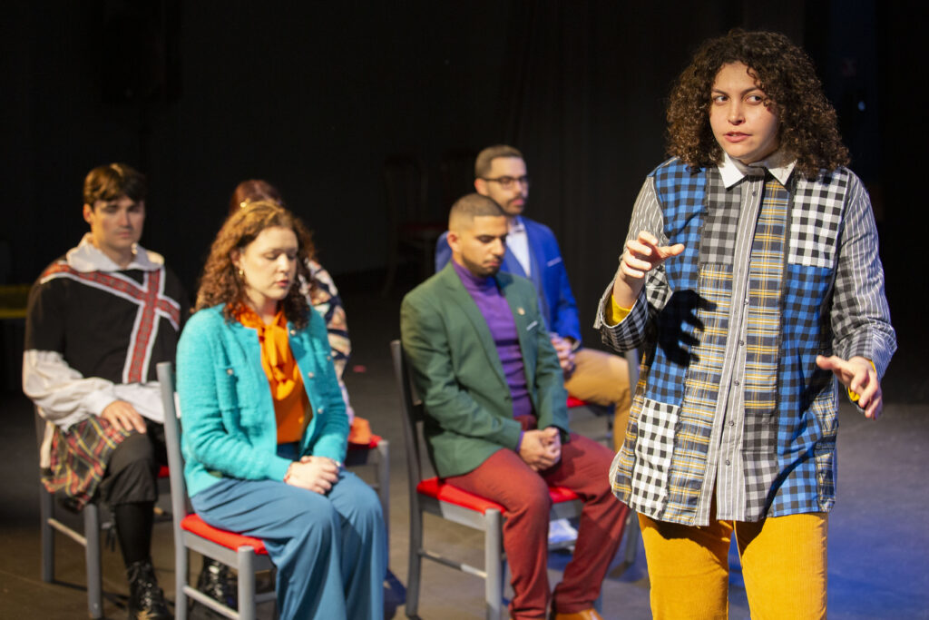 A person with curly brown hair dressed in a multi-patterned shirt and yellow pants, delivering a soliloquy from Hamlet while five other actors in brightly colored street clothes sit on chairs behind them, with their hands clasped in their laps.