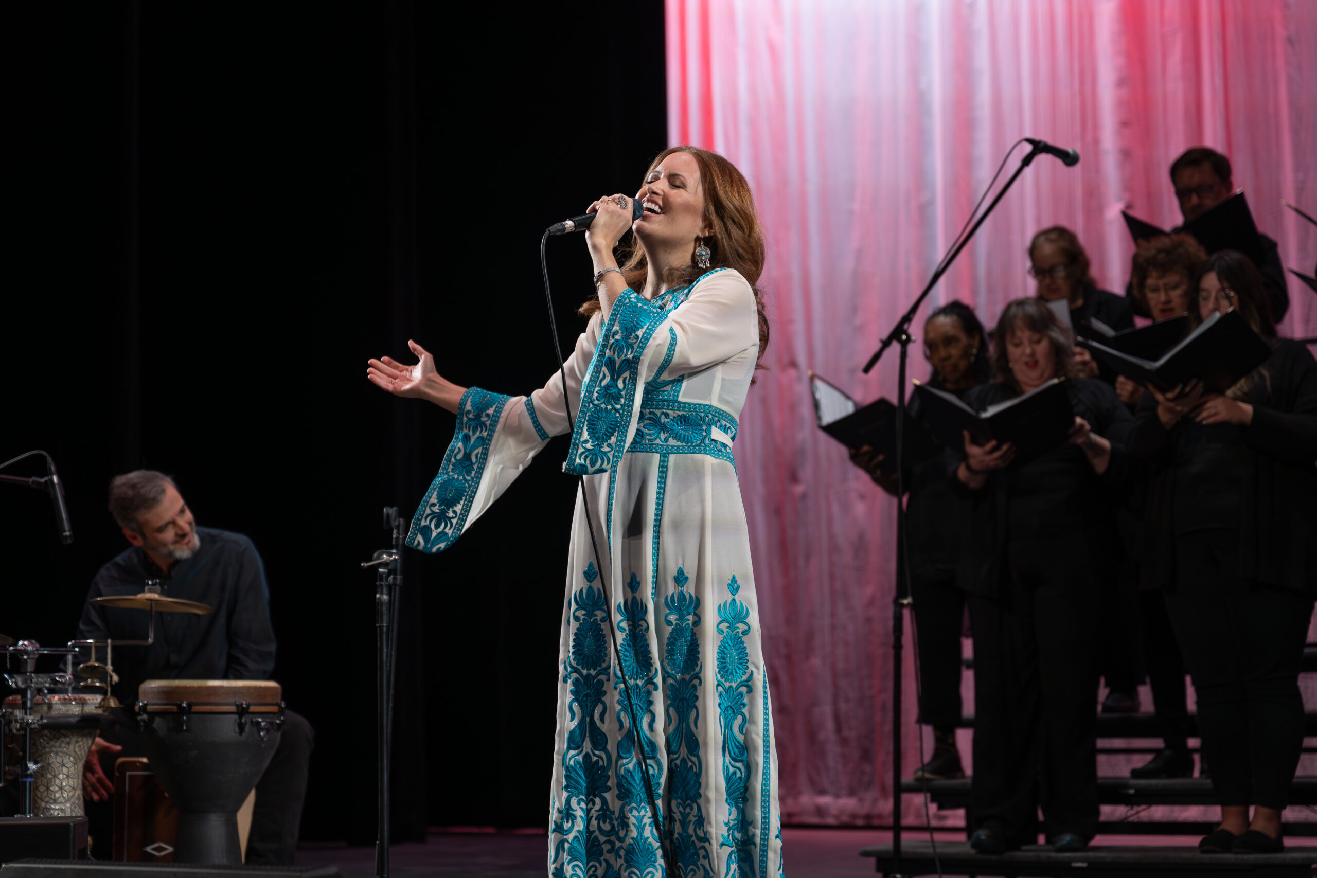 A woman in a long white and teal embroidered dress holds and arm outstretched and sings into a microphone. To her right, a man sits at a drum set, and to her right, a choir stands on rafters singing from black folders.
