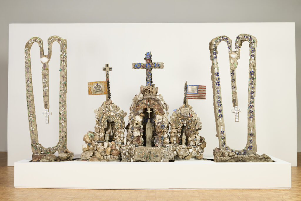 Three ornate sculptures made from small stones. In the center are three arches made from stones with religious statues in the center of each. To each side are identical pieces in a droopy heart shape with mosaic tiles and Christian crosses hanging in the center.