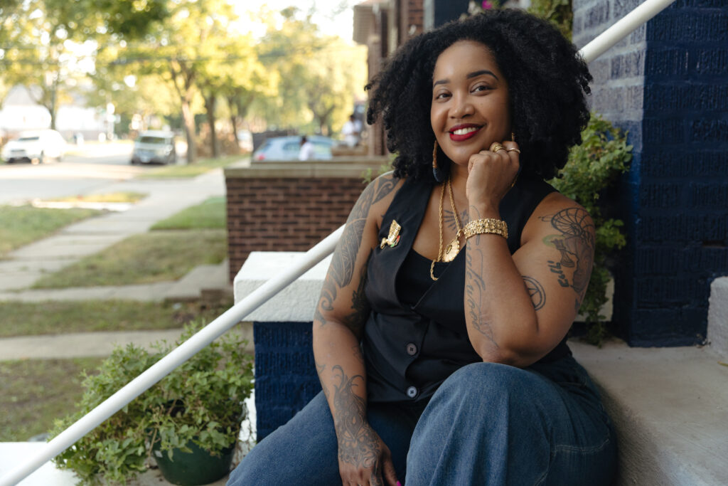 Tonika Lewis Johnson is smiling, sitting on outdoor steps, wearing a black vest, jeans, and gold jewelry, with her hand up to her chin. The background features trees and a sidewalk on a sunny day.