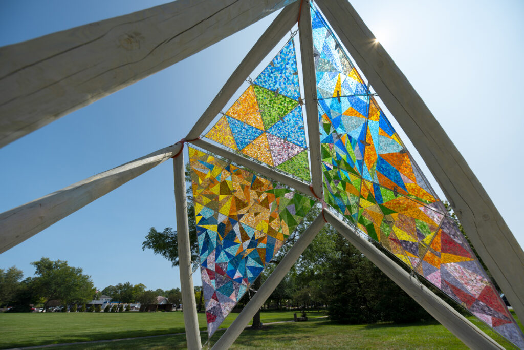 The inside of an open pavilion made of wooden beams supporting brightly-colored glass mosaics in a public park, with sunlight streaming through the colorful glass panels.