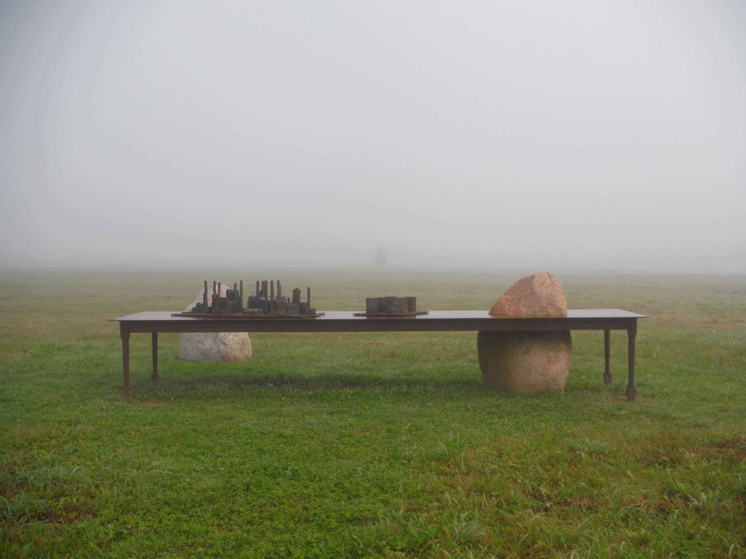 A large sculptural artwork placed outdoors in a foggy field. It depicts a long table with other sculptural elements. There is also a large boulder sticking out through a section of the table.