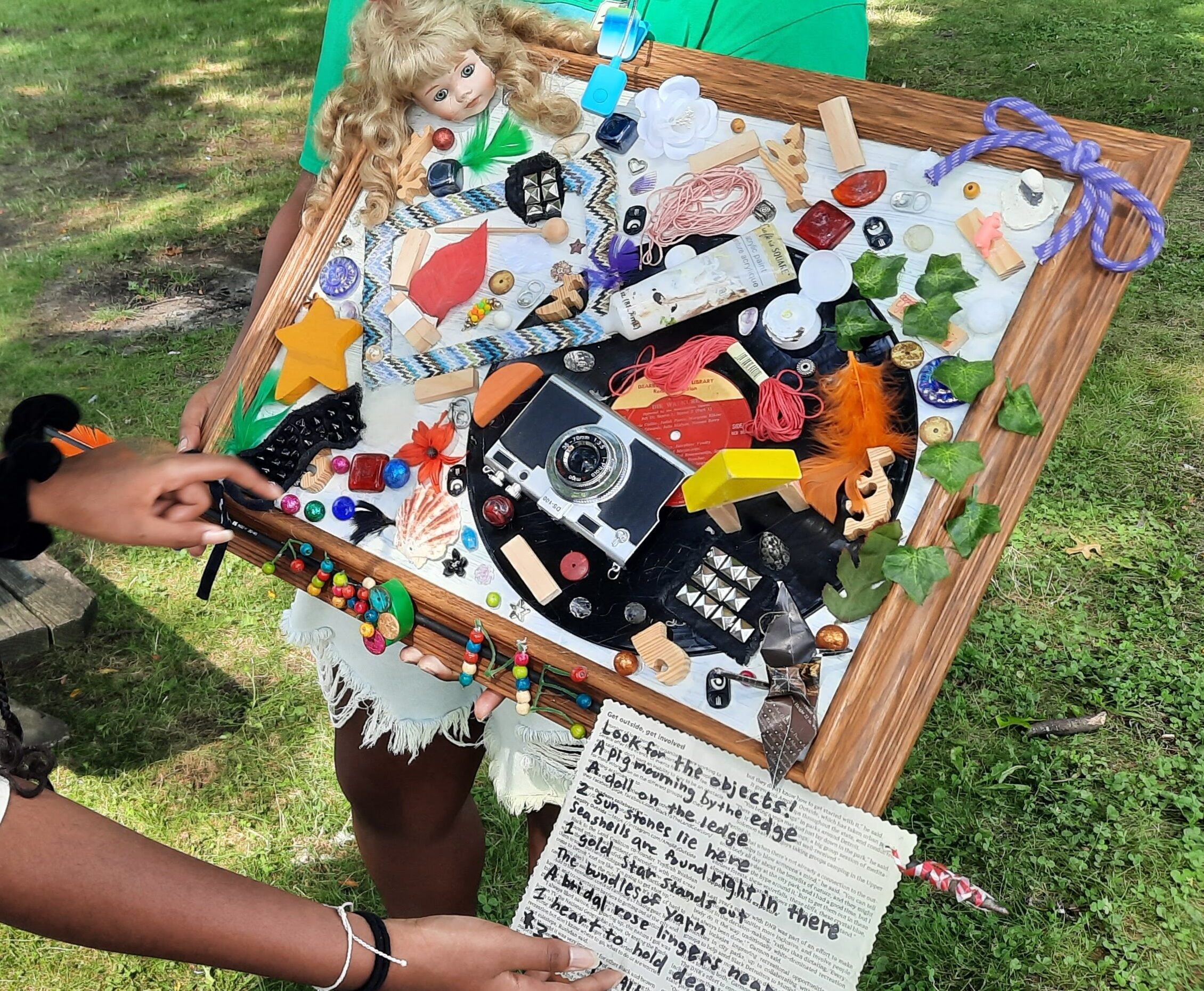 A dark skinned hand pointing to a frame with items glued to it next to a poem.
