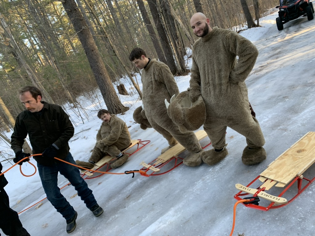 Crew members and people in beaver suits stand beside wooden sleds on an icy forest set.