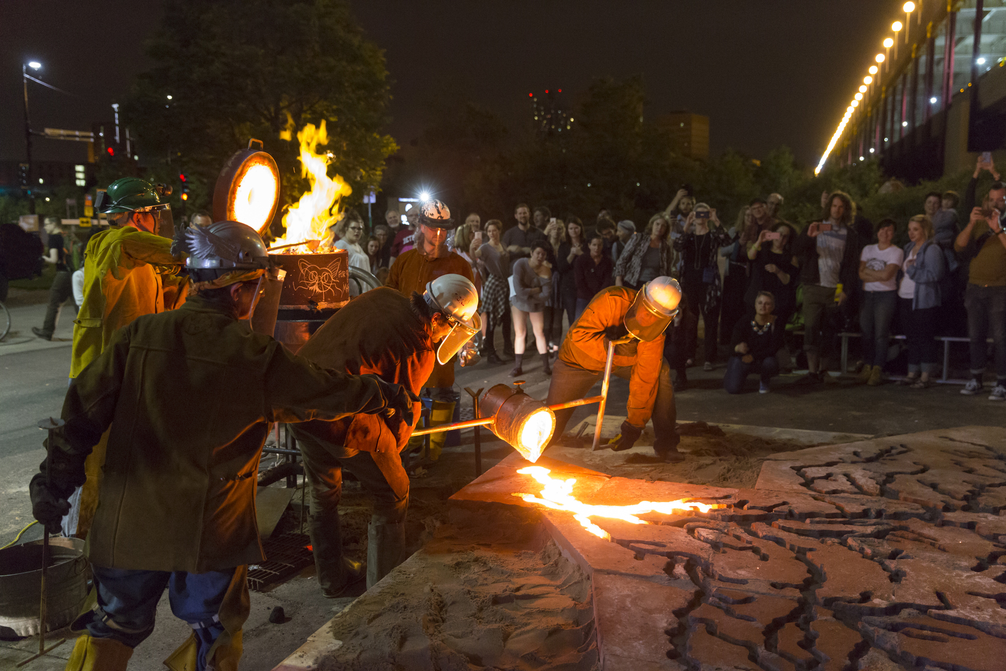 A group of metal workers working next to an outdoor furnace and two of them pour hot metal into a large mold.