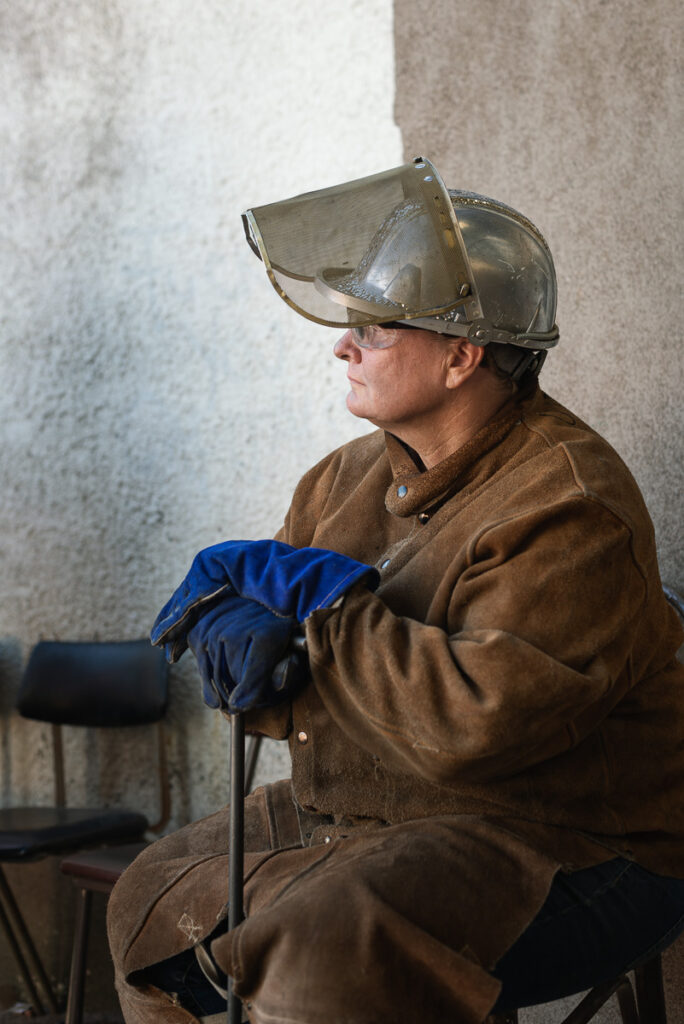 A person sitting and looking away. They are wearing thick brown clothing, blue gloves, and protective helmet for working with hot metals.