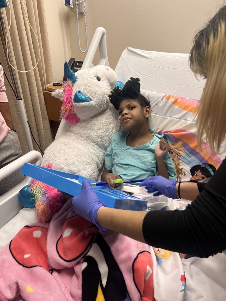 A young child in a hospital bed leaning on a giant stuffed unicorn pillow holding a paintbrush while an adult in blue hospital gloves holds a blue canvas.