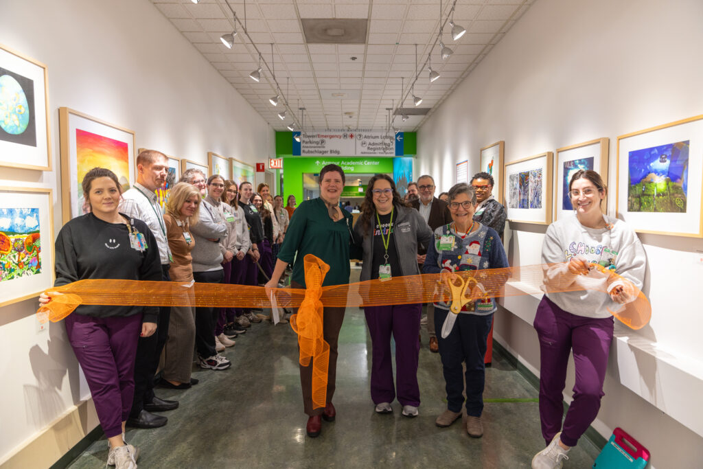 A large group of adult staff in purple scrubs standing in a hospital hallway lined with bright artwork in frames, ready to cut a large orange ribbon.
