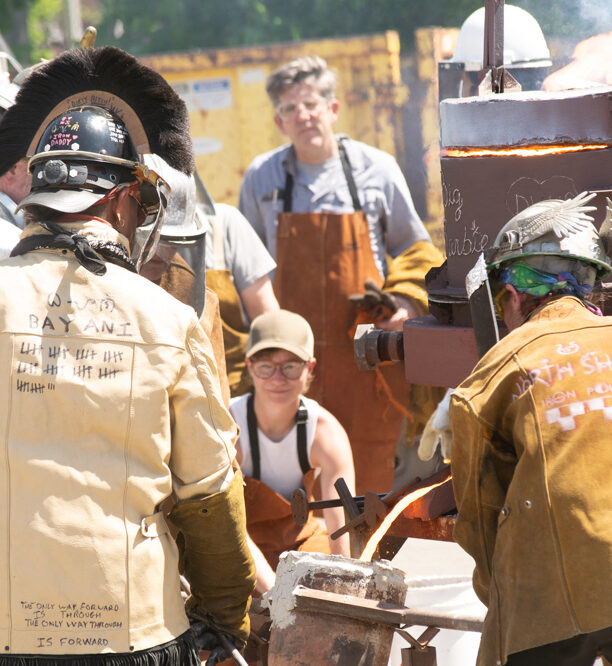 A group of people standing outdoors wearing heavy metal casting protective gear next to a portable furnace.