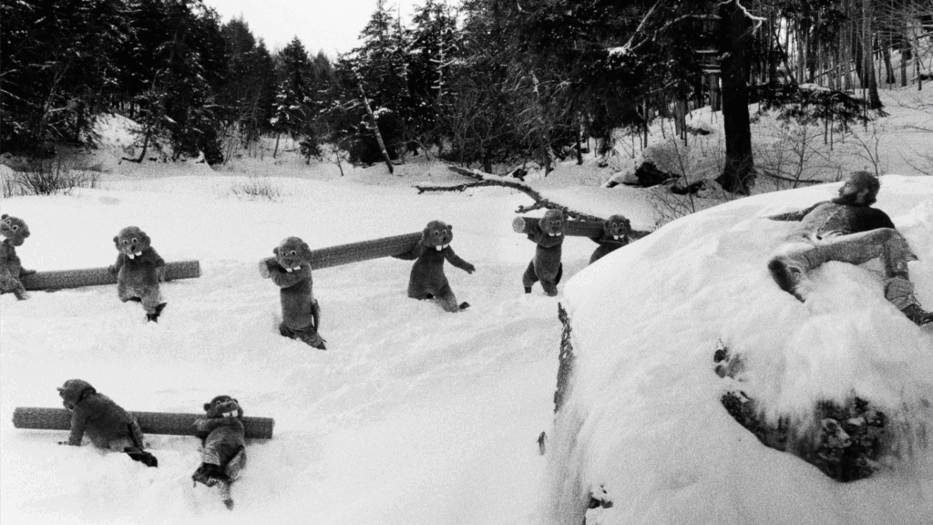 People in beaver suits carry large logs through deep snow in a snowy forest.