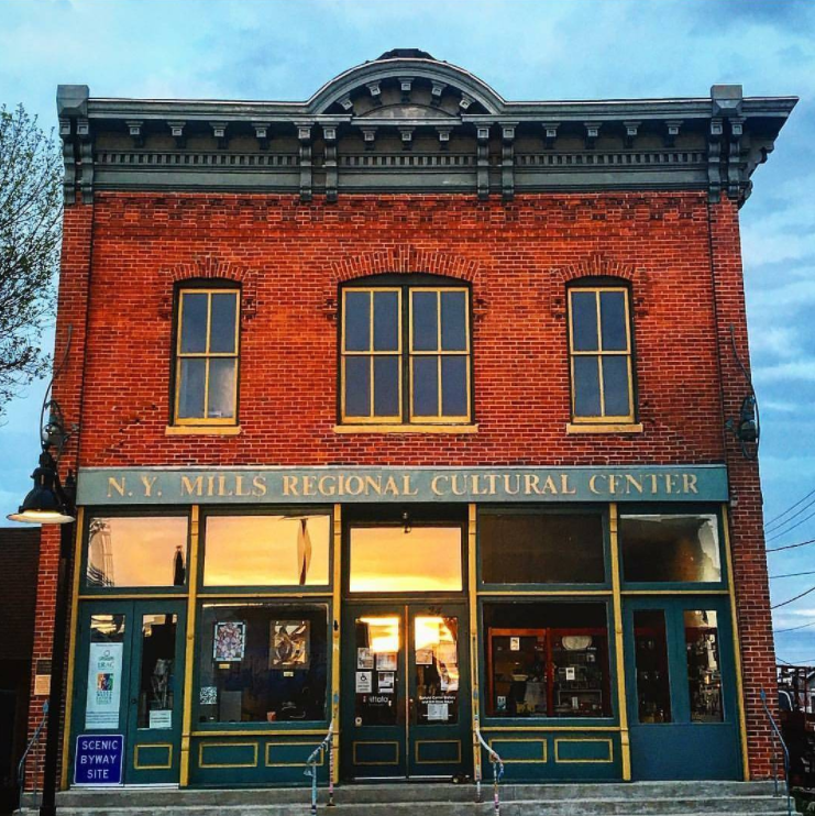 The historic brick facade of the N.Y. Mills Regional Cultural Center in Minnesota. The two-story building has tall windows, green trim, and a sign above the entrance reading “N.Y. Mills Regional Cultural Center.” The evening light glows warmly through the lower windows.
