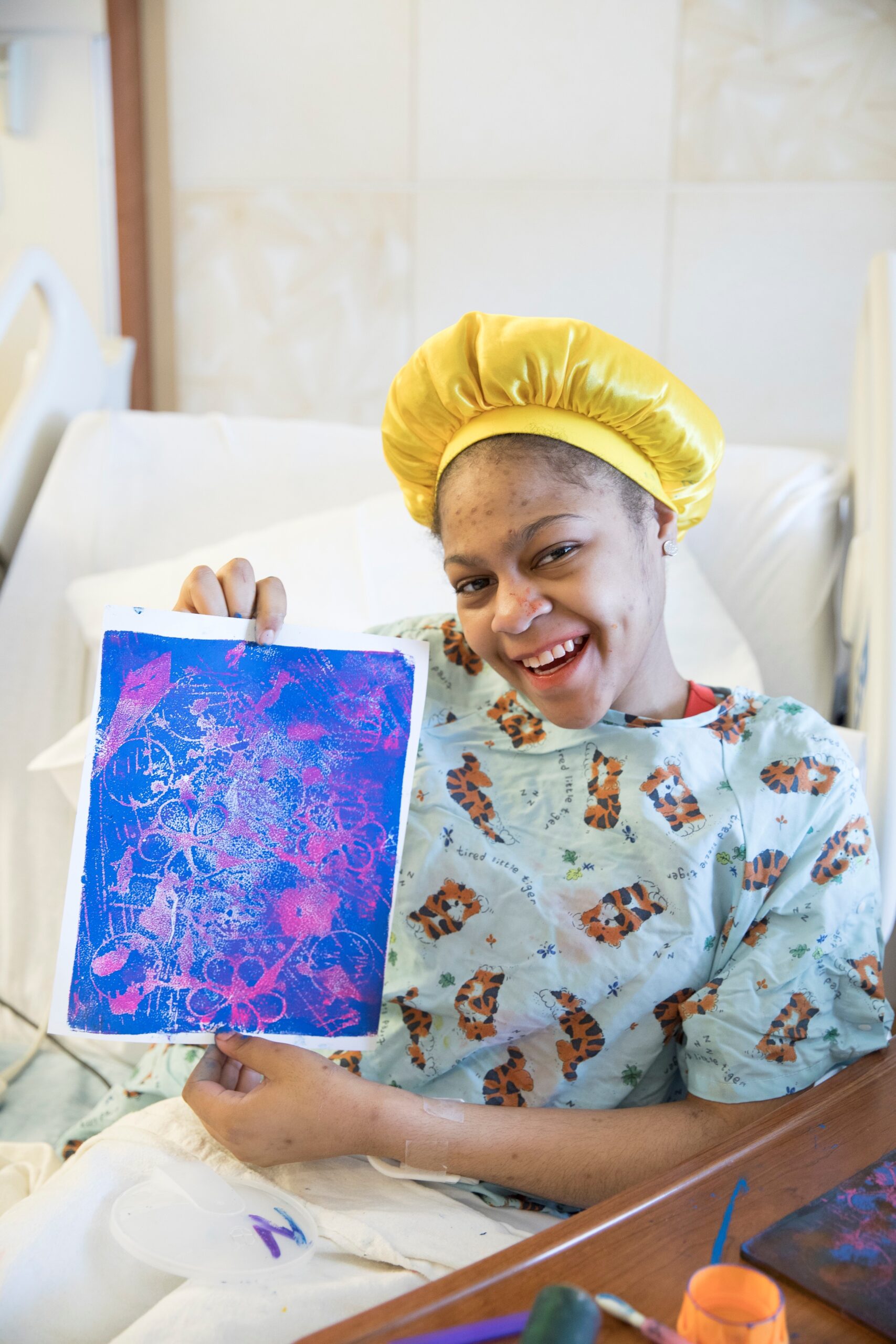 A young patient in a yellow hair bonnet and blue hospital gown with tigers on it, smiling and holding up an original art piece in blues, yellows, and purples from her hospital bed.