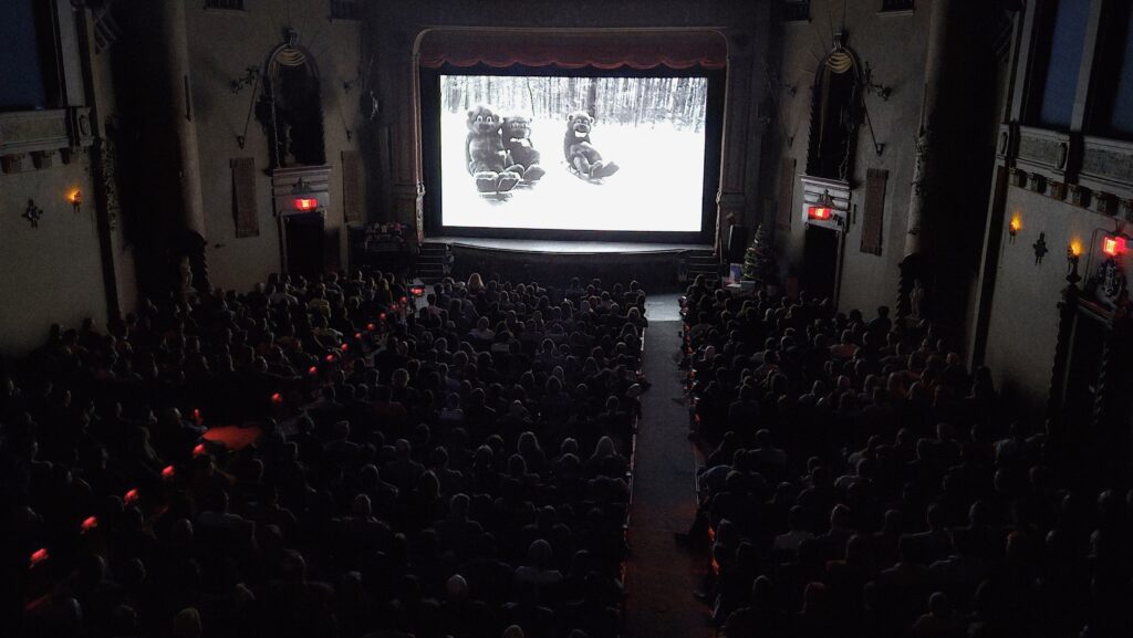 A full audience watches a black-and-white film showing people in beaver suits on a snowy trail.