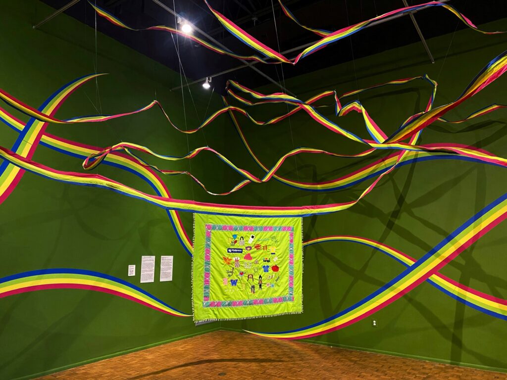 An artwork suspended in the middle of a room with green walls. It is a bright green textile artwork with embroidery. There are also wave-like fabric rainbows suspended around the piece.