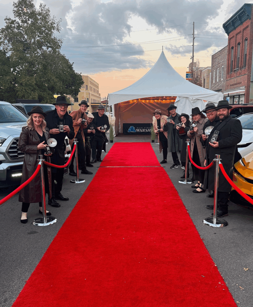 People standing with cameras next to a red carpet in a perking lot.