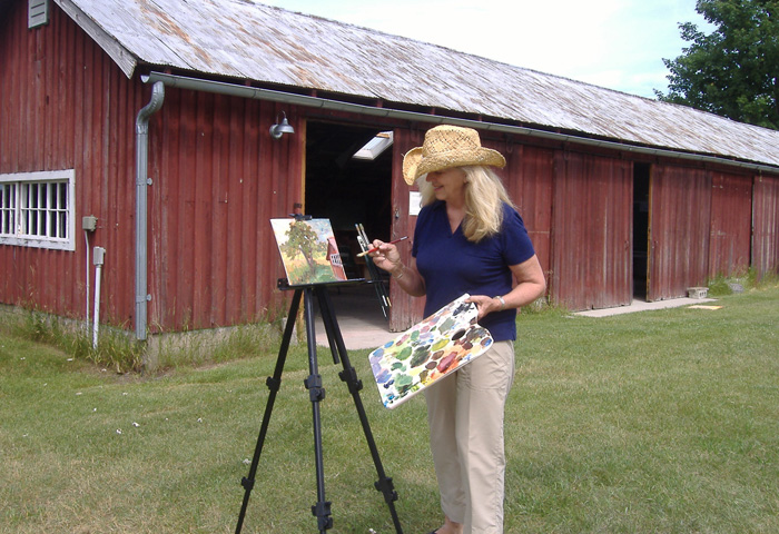 A woman with a cowboy hat and paintbrush outside a red barn.