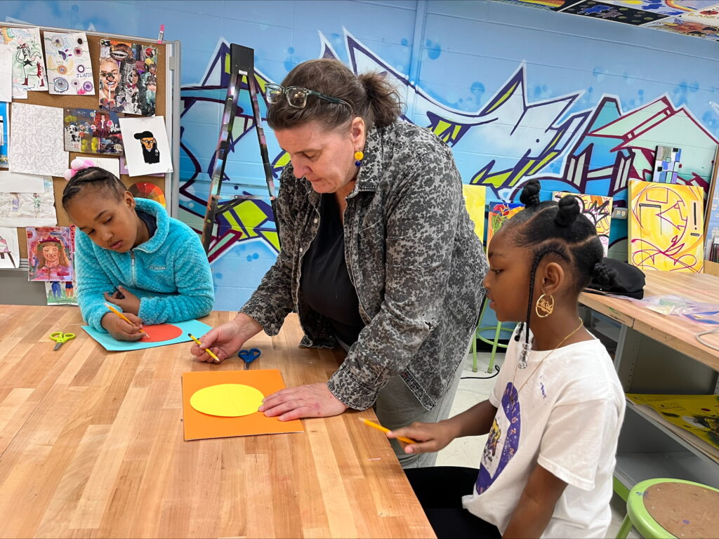 A person hunching over a table in a classroom as they demonstrate to two young people who are sitting at the table and observing.