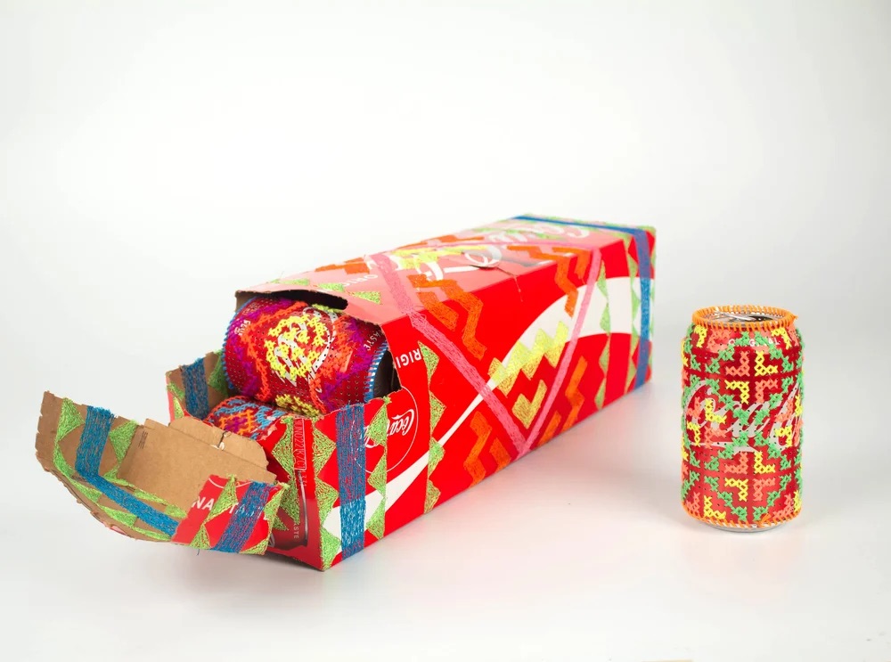 An opened box of soda with one can right next to it. The box and the cans are embroidered with thread depicting a repeating geometric pattern