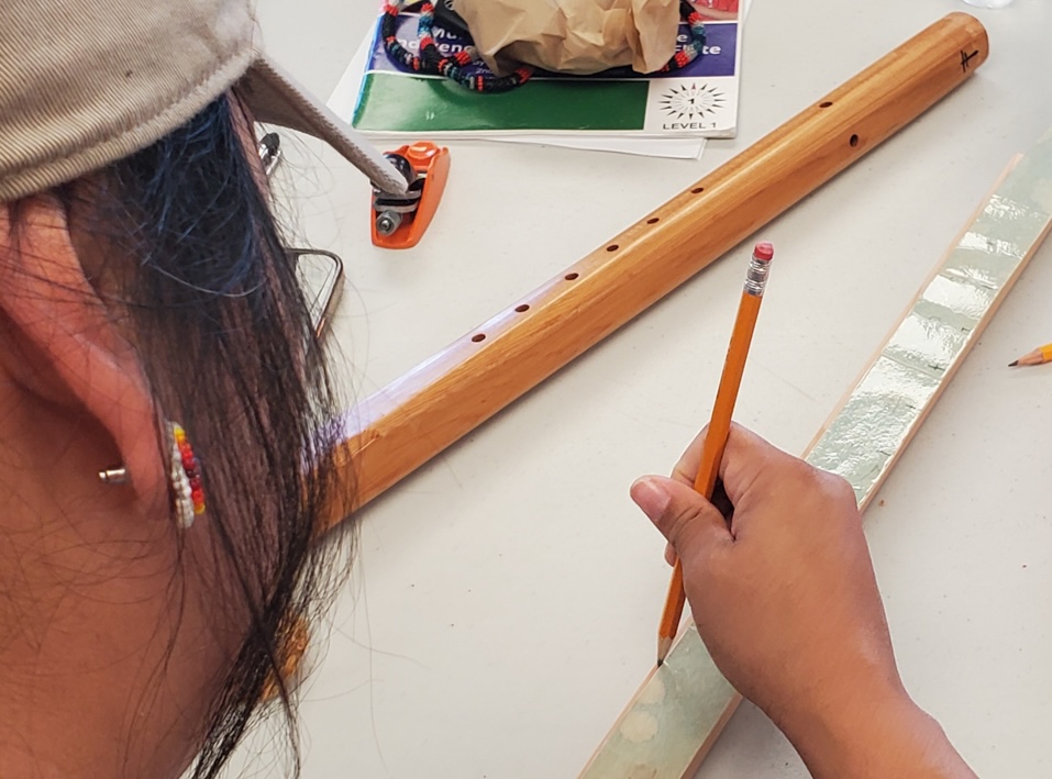A person at a table using a pencil on a piece of wood as a handmade wooden flute rests next to them.