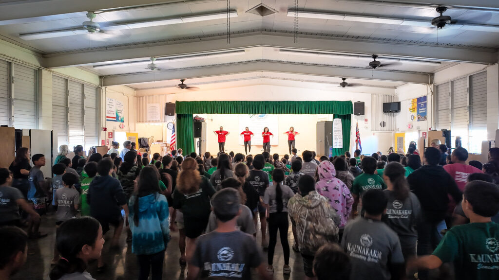 Four percussive dancers in red t-shirts performing on a small stage surrounded by a green proscenium curtain in a large open room filled with young students imitating the same moves.