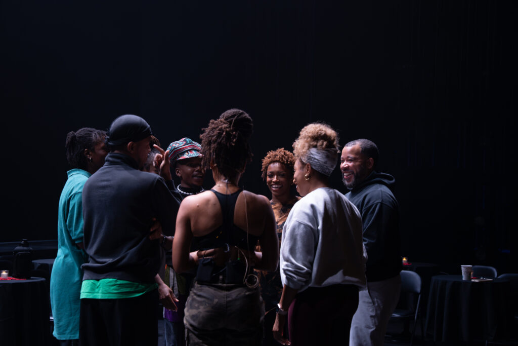 A circle of adult performers facing each other and laughing, with choreographer Ryan K. Johnson at one end in a black hoodie.