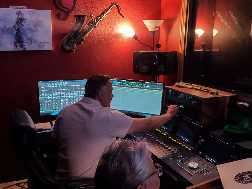 A person with a white polo shirt facing a table with multiple computer screens and audio recording equipment. Their arm is reaching for an equipment in front of them.