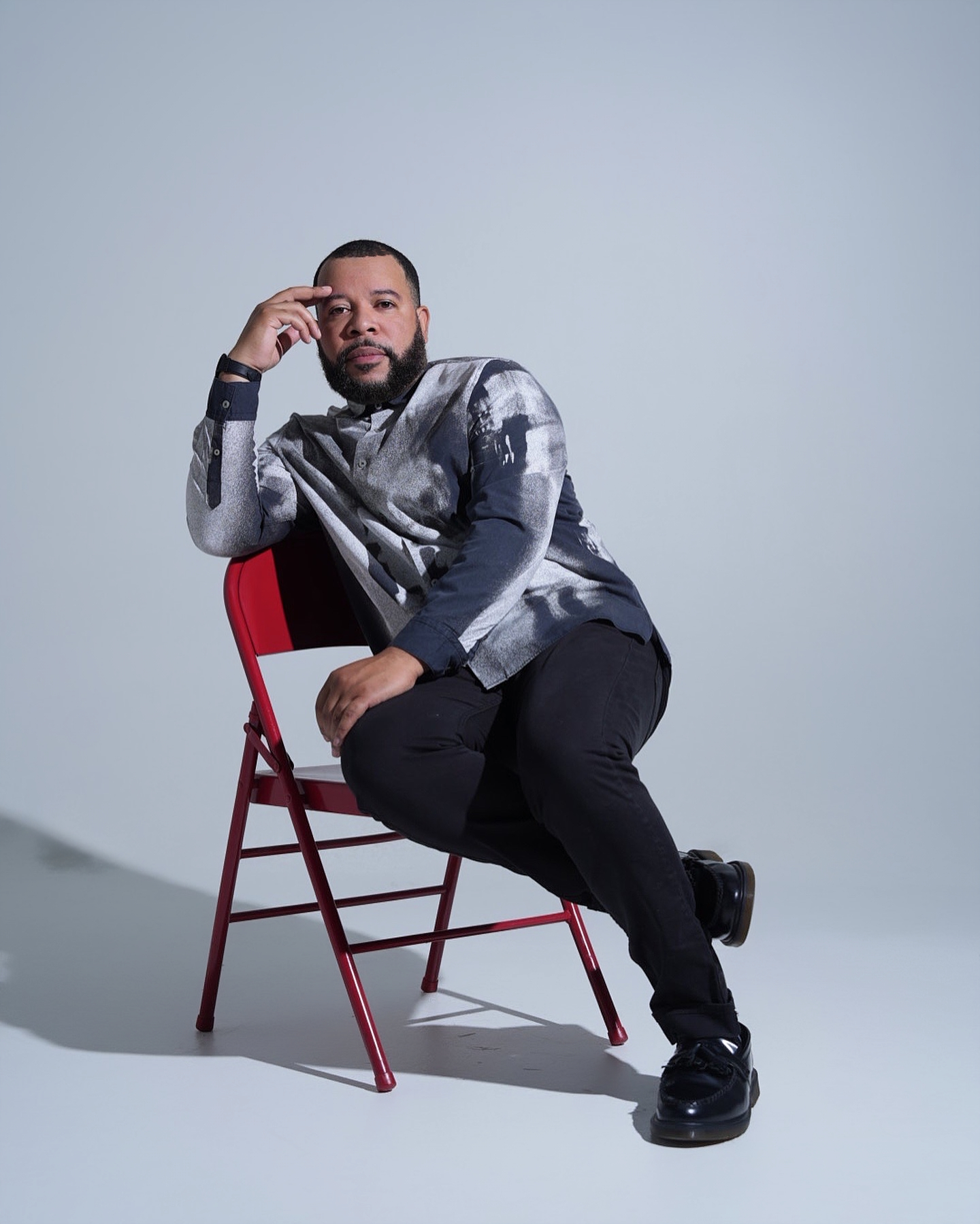 A middle-aged African American man with a full beard and short black hair sitting on a red chair, with shiny black shoes and two fingers on his temple.