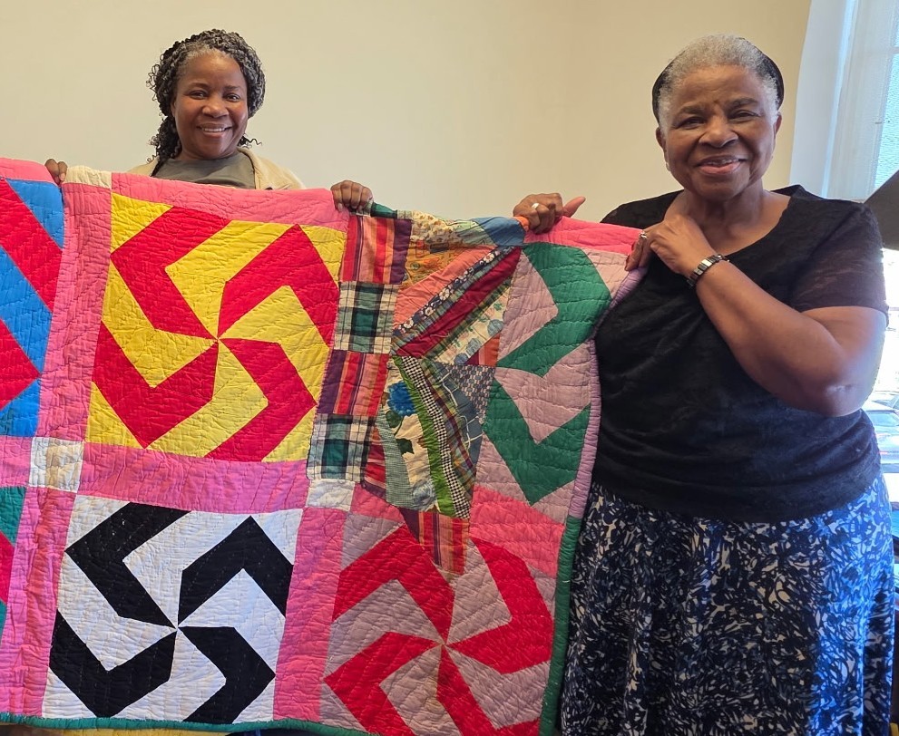 Two women with dark skin tones holding up a colorful quilt.