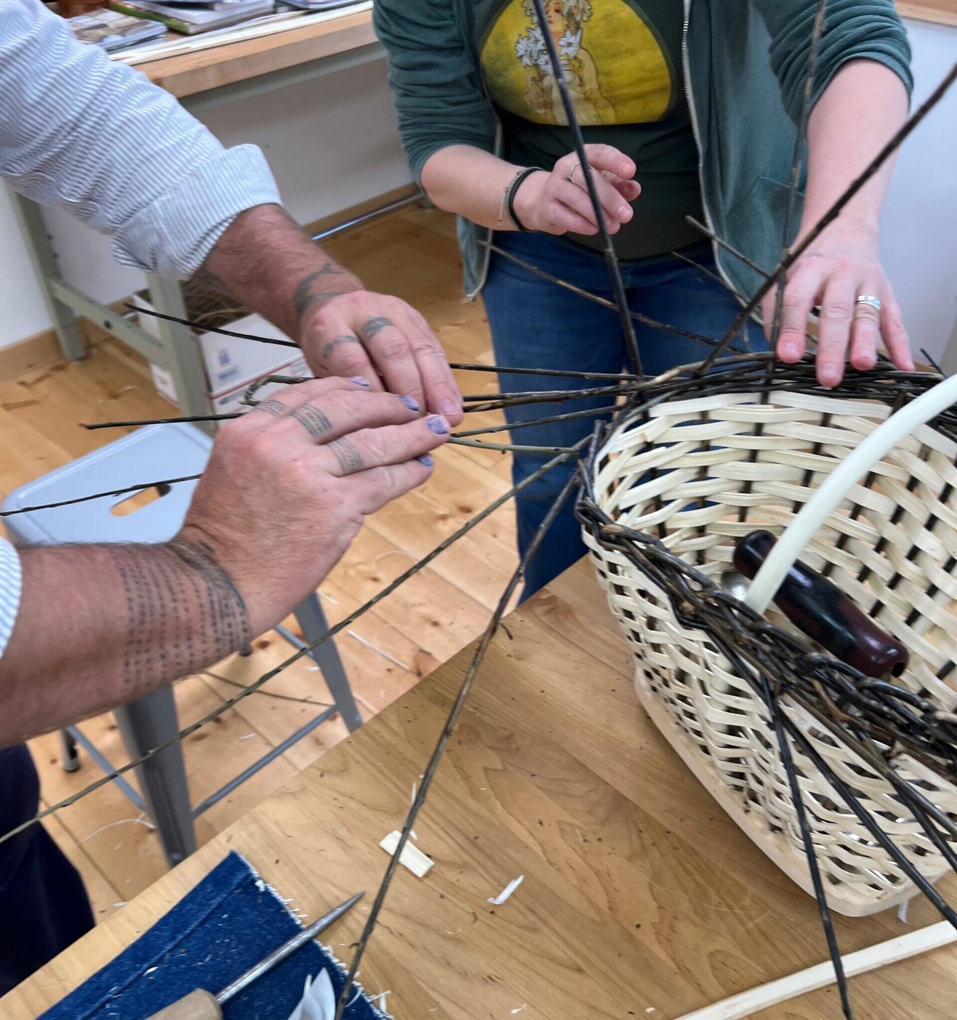 Two sets of hands weaving a white and brown basket on a wooden workshop table.
