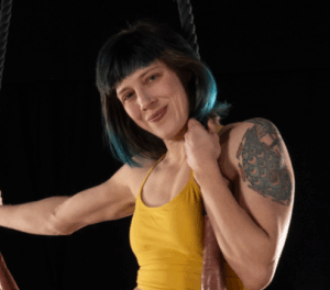A person with blue-tinged, shoulder-length hair, a colorful tattoo on their left shoulder, and a yellow top, sitting on a trapeze.