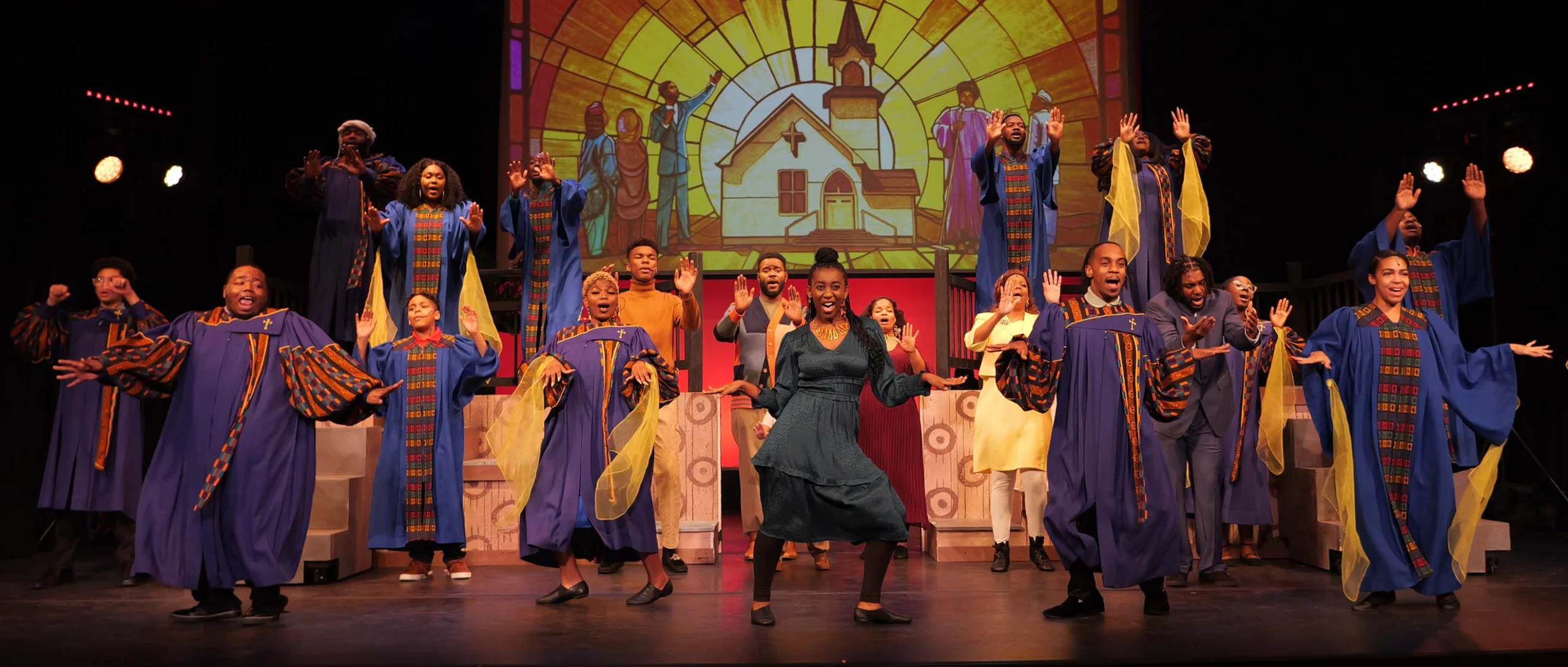A large ensemble performs onstage in colorful choir robes, singing and dancing with raised hands in front of a stained-glass church backdrop