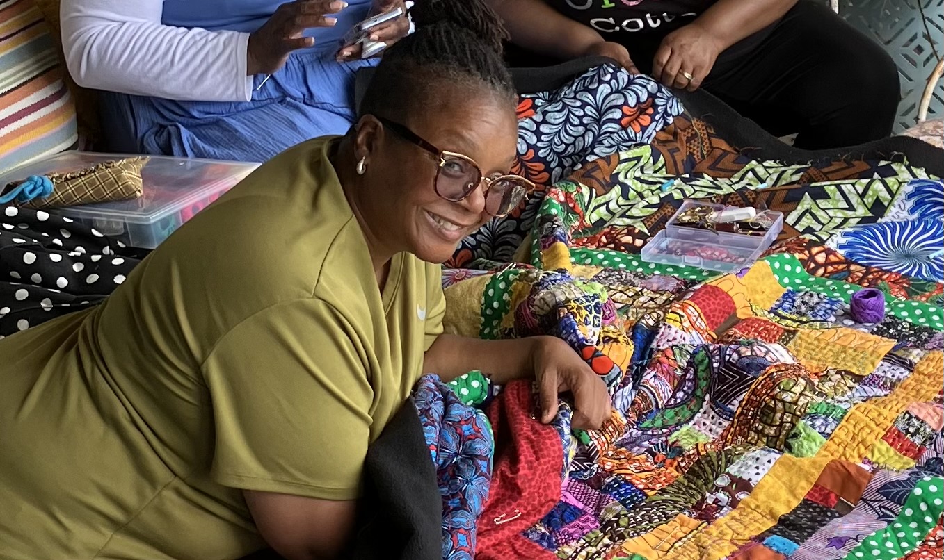 A woman with dark skin smiling and lying on the ground on a quilt.