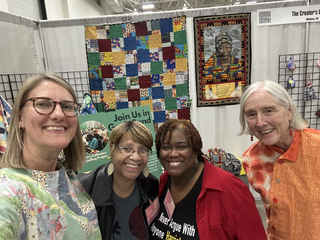 Three women smiling in front of a quilt display indoors.