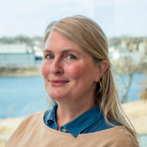 A woman with light skin, blonde hair, blue-gray eyes. and gold hoop earrings smiling.
