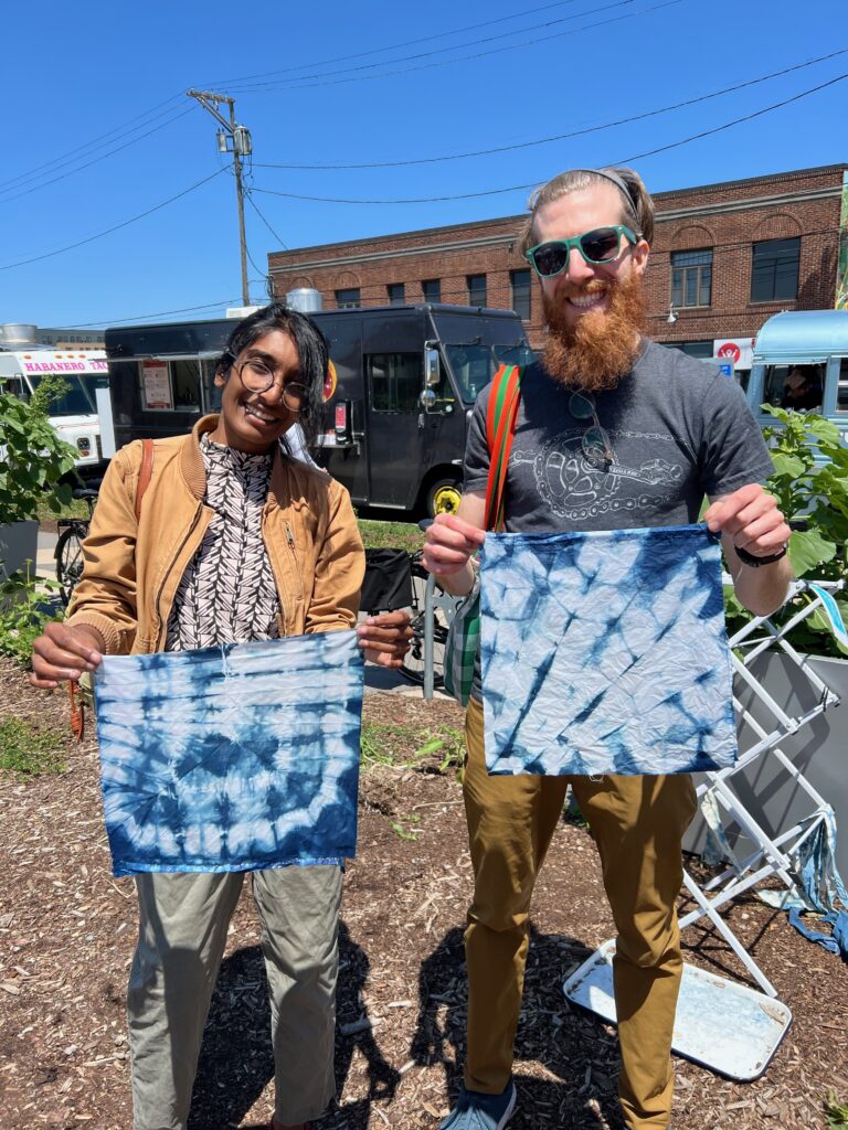 Two people smiling and standing outside, holding up squares of fabric that has been tie-dyed a deep blue color.