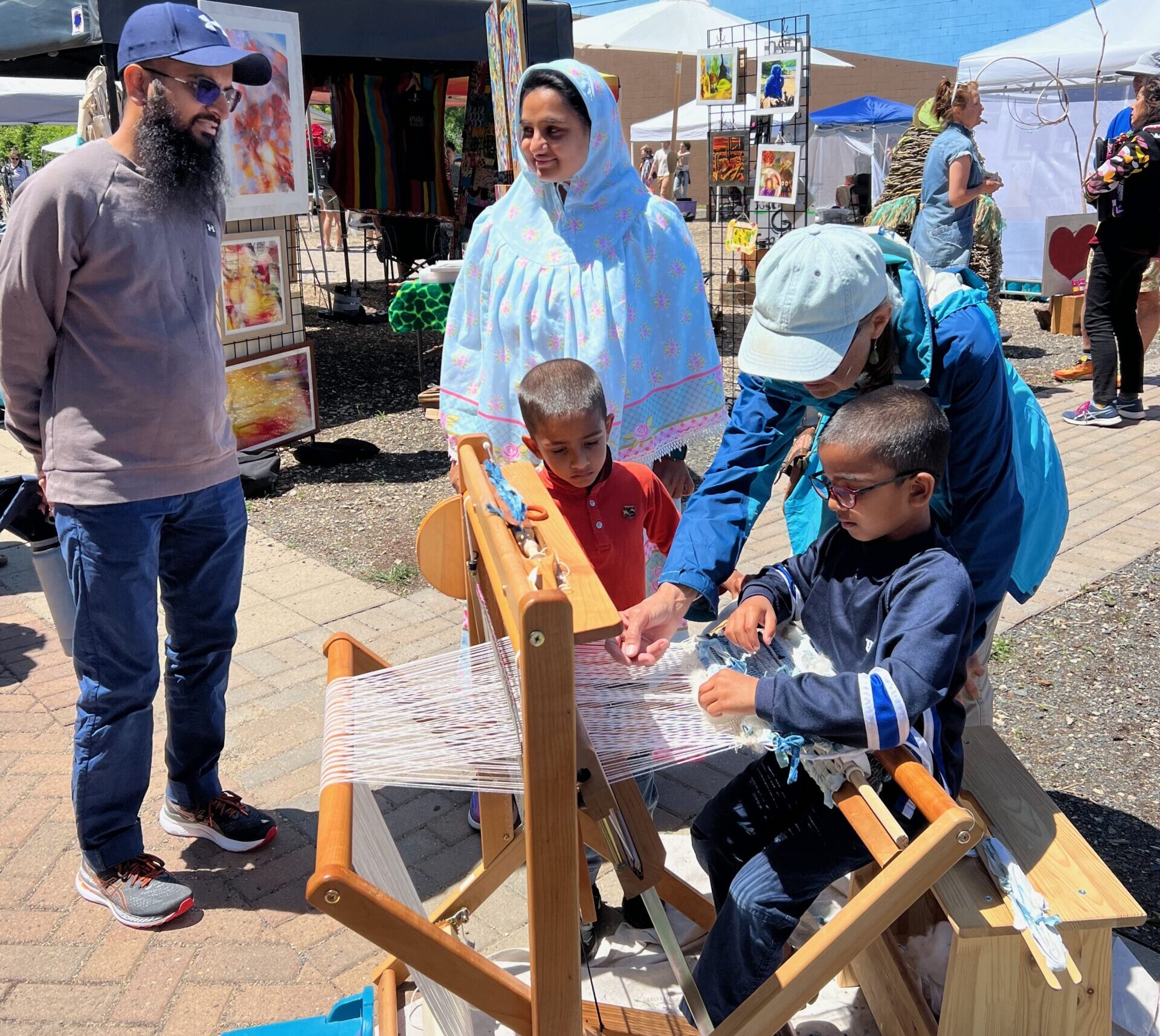 A young child sitting outside at a wooden loom strung with white strands, with an older adult leaning next to him to demonstrate and another young child watching. Two adults stand nearby to watch.