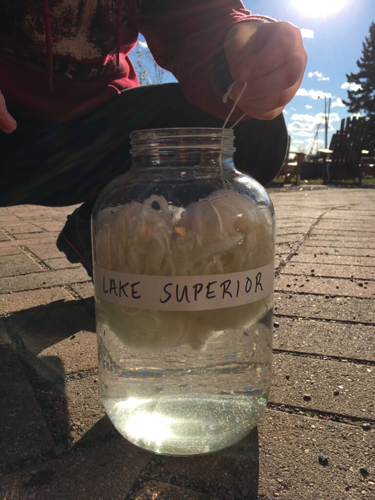 A glass jar labeled "LAKE SUPERIOR" filled with water and a ball of white fibers. A person crouches behind holding the ends of two of the white fibers out of the top of the jar.