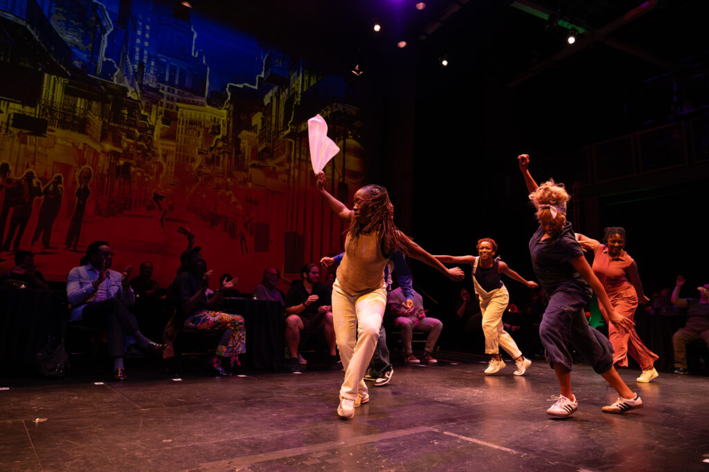 Five Black percussive dancers performing on a black box stage surrounded by seated audience members and a projected illustration of New Orleans filling one the wall.