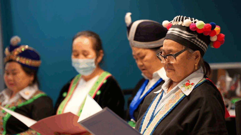 Four older Hmong adults wearing colorful traditional headpieces and black cardigans, holding folders of sheet music.
