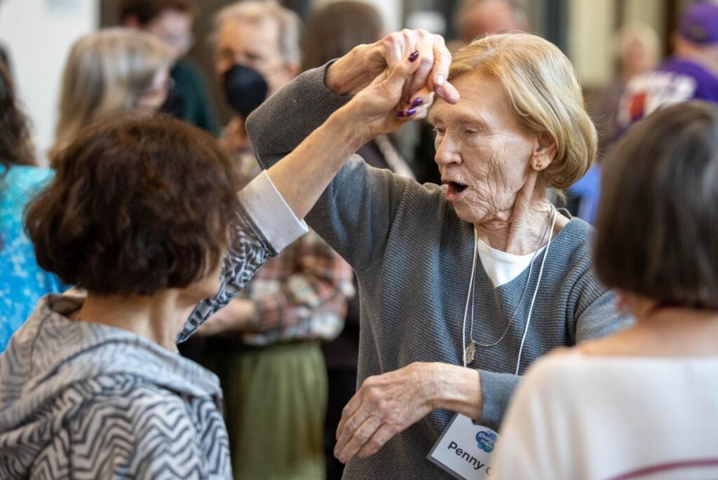An older woman with short hair and a gray sweater twirling another older woman around in a dance, surrounded by other dancers.