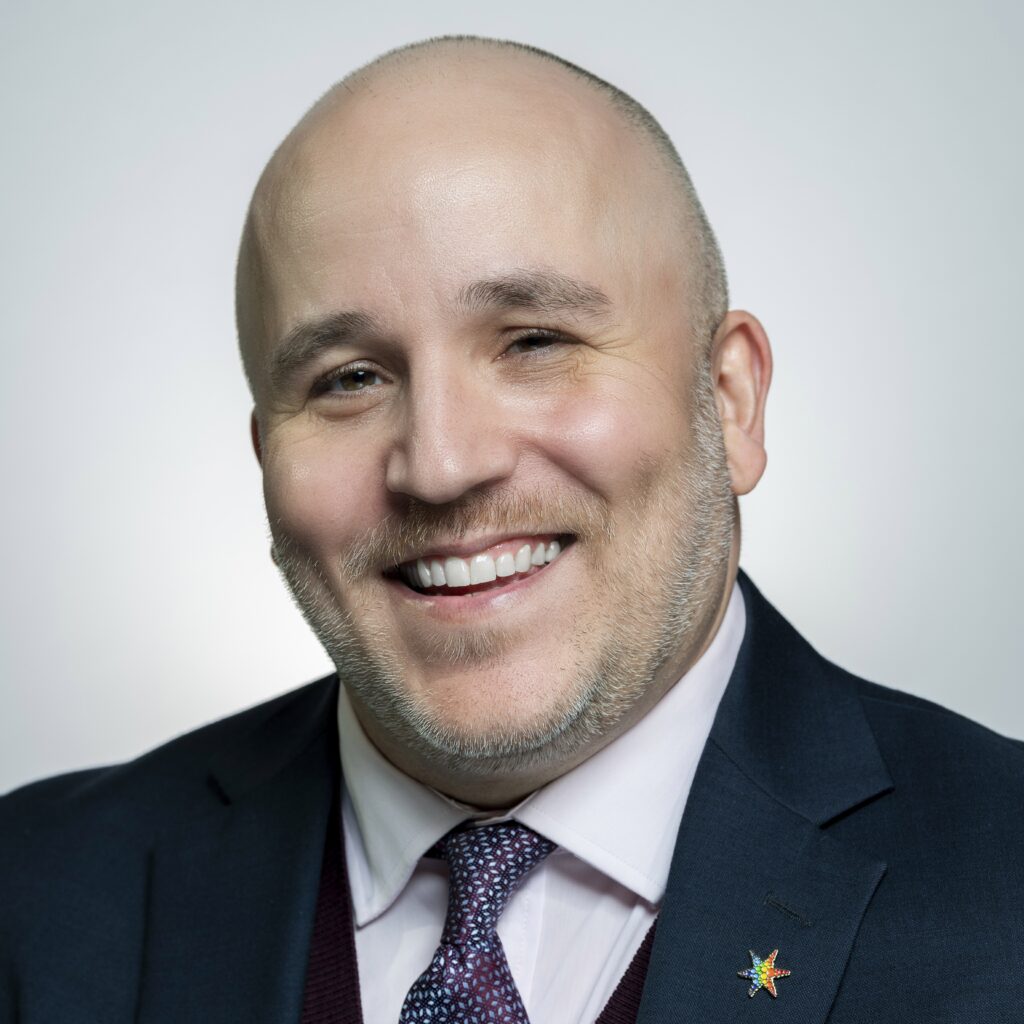 Headshot of a man with light skin tone, wearing a suit and tie, smiling.