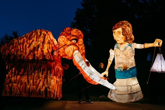A large puppet of a young woman carrying a lantern and leading a woolly mammoth by the trunk.
