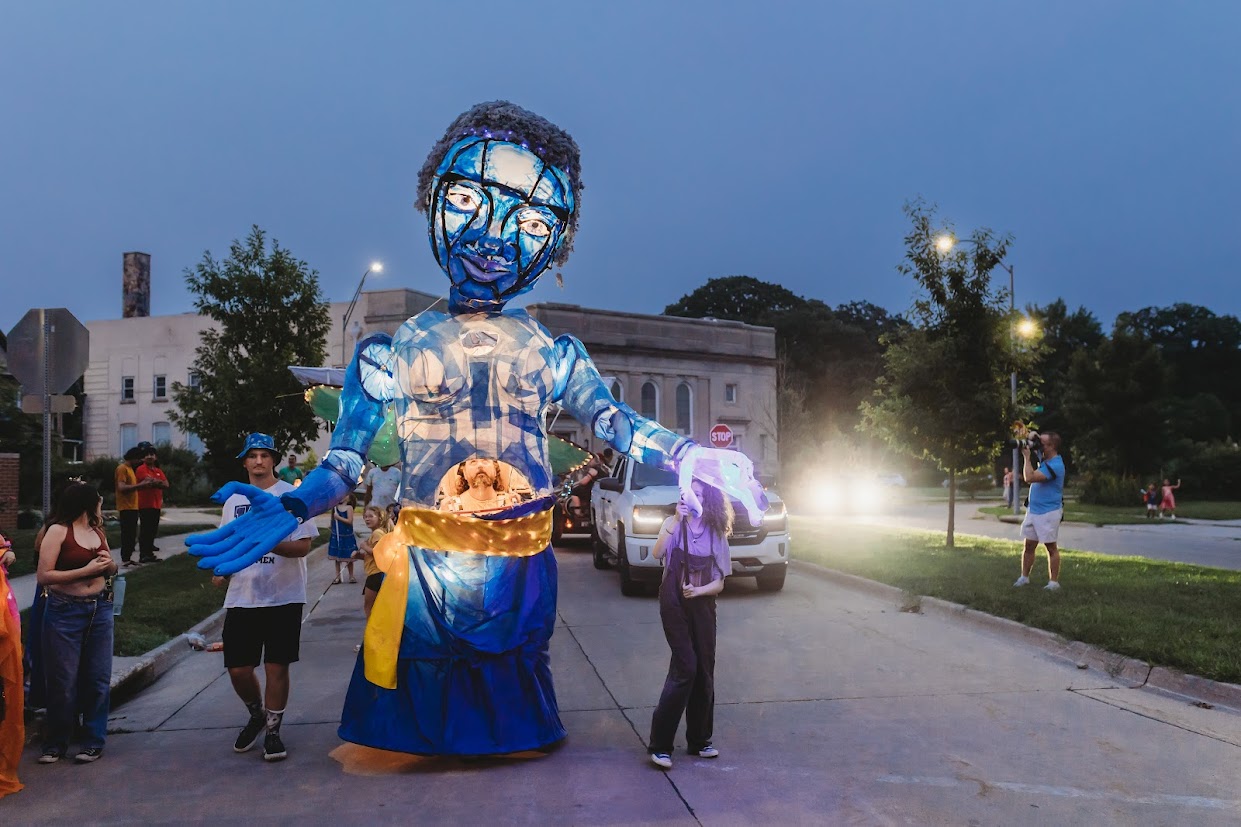 A large puppet - close to 15 feet tall - illuminated by internal lights, being guided by multiple puppeteers. It is in the shape of a human with blue skin wearing a wrapped blue skirt and gold sash.