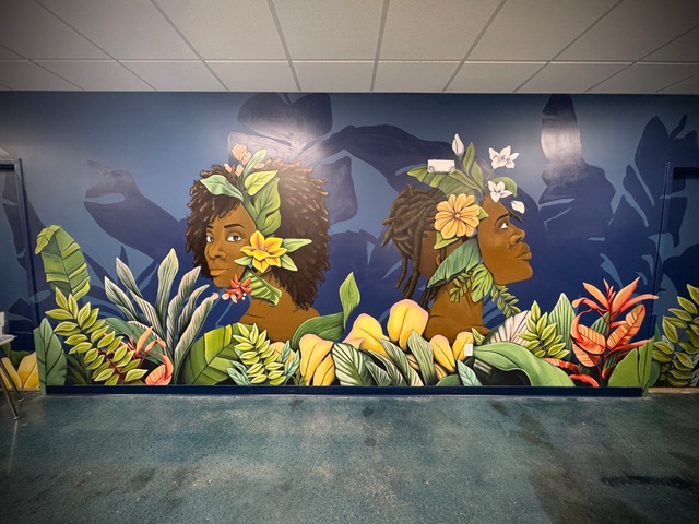 A wall mural featuring portraits of two Black individuals with flowers and vines in their hair, above a bed of plants in various colors, shapes, and textures.