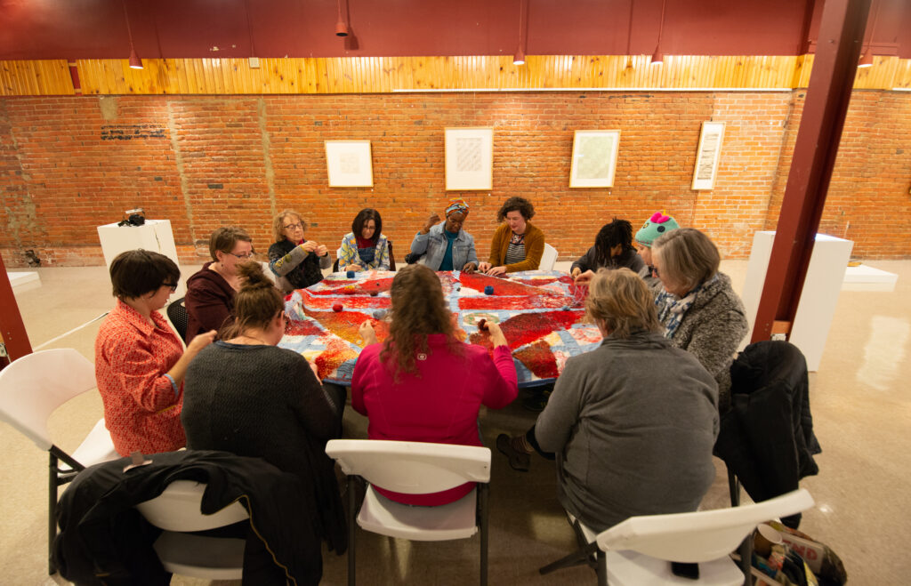 A group of adults sitting at a round table, stitching a large multicolored cloth.