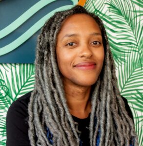 Headshot of a smiling person with medium skin tone and long gray locs, wearing a black top. The background features a vibrant tropical pattern with green palm leaves and abstract blue and orange shapes.
