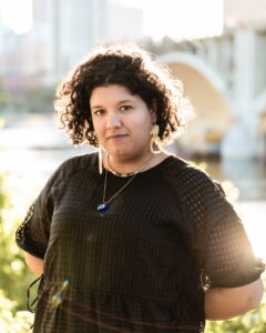 Medium close shot of a person with curly, shoulder-length dark brown hair wearing a black dress and white earrings.