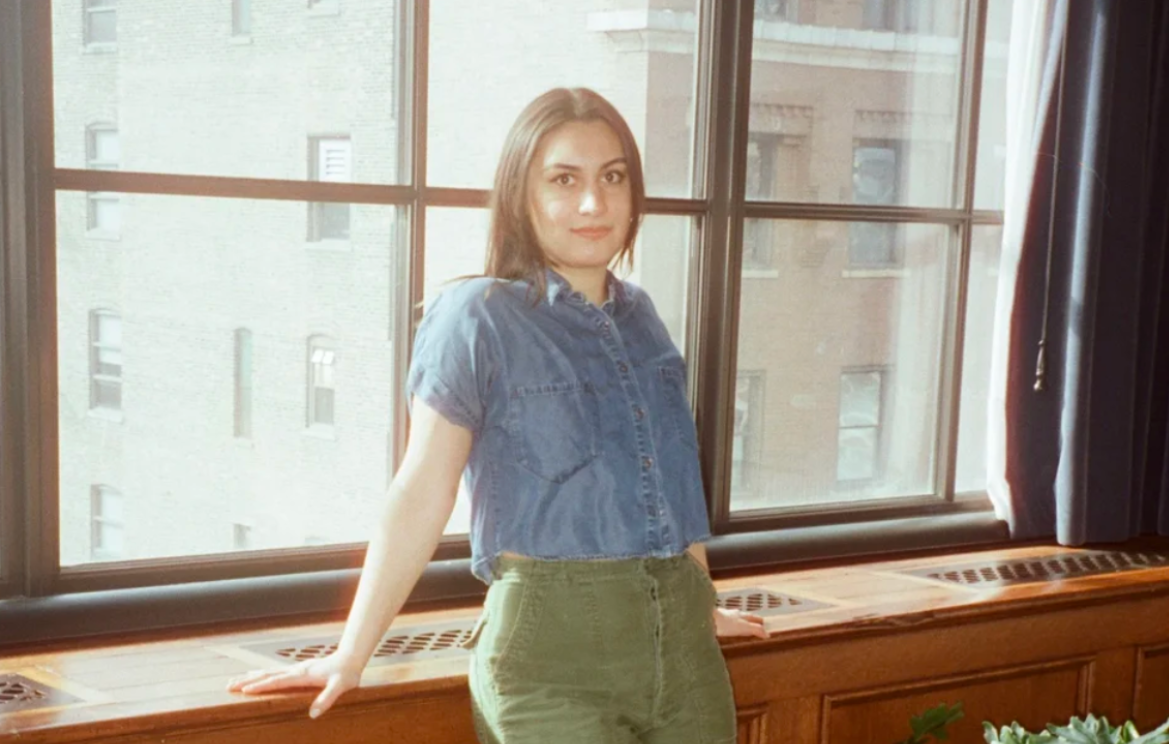 A woman with brown long hair and medium skin tone poses near a window.