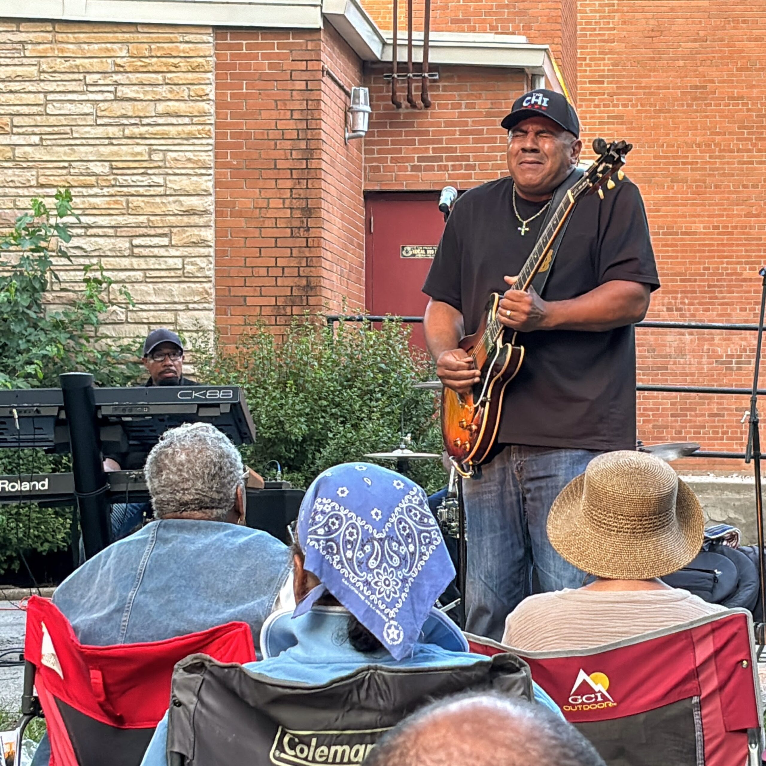 A blues guitarist performs outdoors in front of a seated audience. The musician stands on a small stage area while audience members watch from folding chairs, with a keyboard player visible behind him.