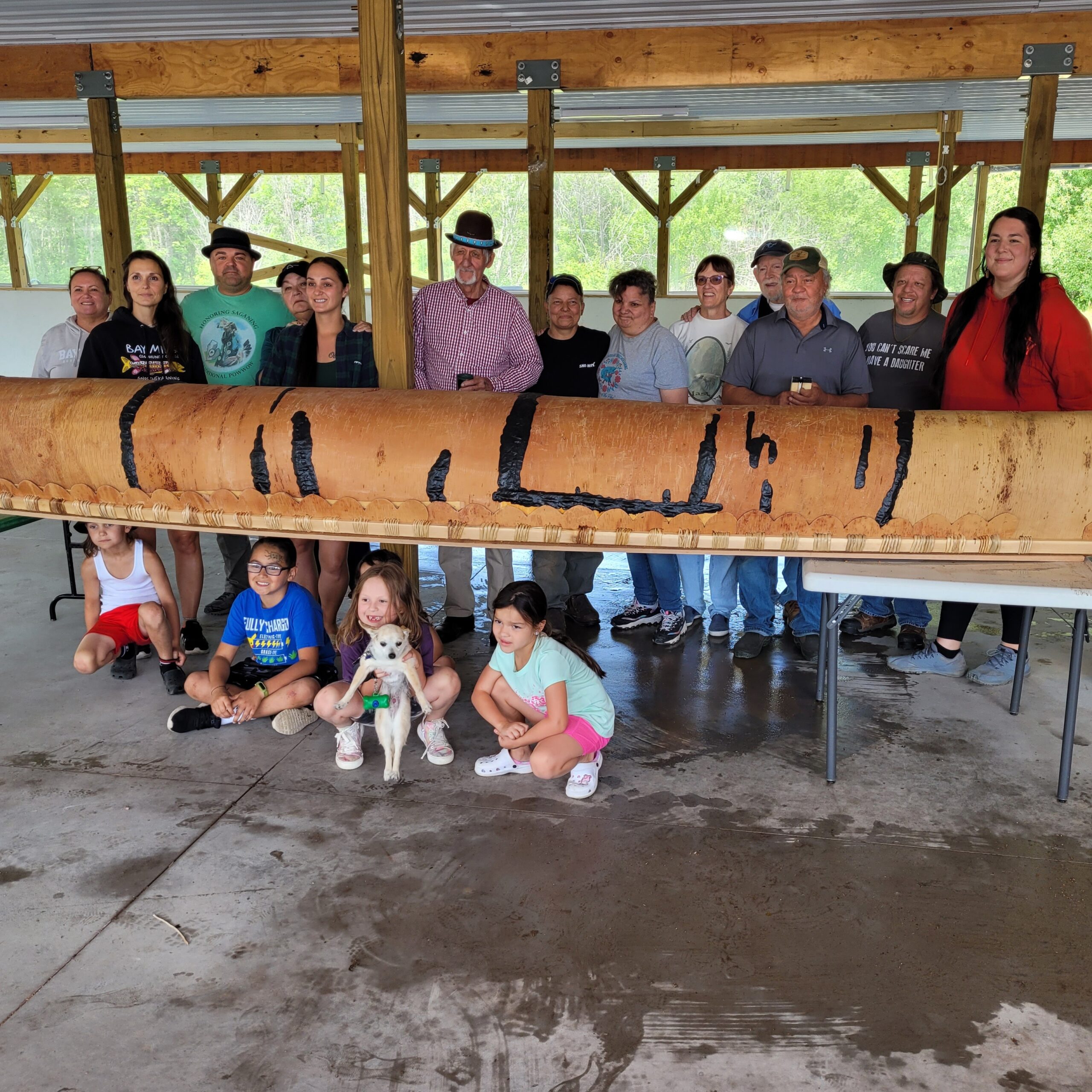 A large group of adults and children stand behind a long wooden birchbark canoe under an open-sided shelter. The canoe rests on tables, and several children sit or kneel in front. Trees are visible beyond the structure.